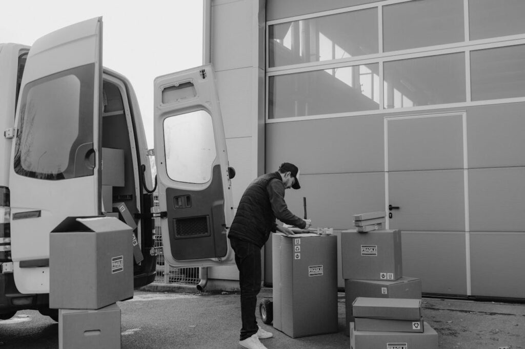 Monochrome image of a courier managing packages outside a warehouse, illustrating logistics workflow.
